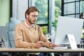 Concentrated young man holds a card in his hands, sits at a desk in the office in front of a computer, makes an online Royalty Free Stock Photo