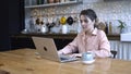Concentrated young woman sitting in the kitchen at wooden table and typing on her personal silver laptop with the cup of Royalty Free Stock Photo