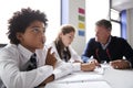 Concentrating Male High School Student Wearing Uniform Working At Table With Teacher Talking To Pupils In Background