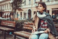 Cute shot of lovely girl sitting on a wooden bench outside, writing a poem, looking lost in thoughts and dreams.