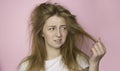 The concept of dry lifeless hair. A woman on a pink background holds her disheveled, tangled hair and looks at it with a