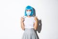 Concept of halloween celebration and covid-19 pandemic. Image of asian girl in blue wig pointing at medical mask