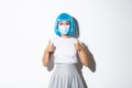 Concept of halloween during covid-19 pandemic. Image of surprised asian woman in blue wig and medical mask showing