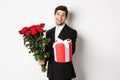 Concept of holidays, relationship and celebration. Image of handsome smiling guy in black suit, holding bouquet of red