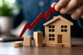 A concept image showing a hand holding a red arrow pointing upwards next to wooden house and bar graph, representing real estate