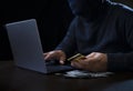 hacker in black hoodie and thief mask using laptop to hack credit card, a lot of money on desk Royalty Free Stock Photo
