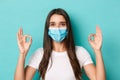 Concept of pandemic, covid-19 and social distancing. Pretty modern girl in white t-shirt and medical mask, showing okay Royalty Free Stock Photo