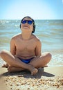 The concept of relaxation and tranquility. A teenager boy sitting on the sand in swimming trunks, blue sun glasses, is engaged in Royalty Free Stock Photo