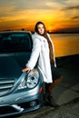 Strong independent young woman with car. Beautiful serious girl fashionably stand near vehicle at sunset