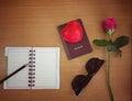 Concept travel valentine plan : pen on open note book, passport, red heart plastic, pink rose on wood table