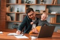 Conception of multitasks. Businessman in formal clothes is working indoors. With toddler Royalty Free Stock Photo