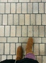 Female feet wearing brown boots, purple tone pants and black coat standing on old stone pavement with straight lines pattern Royalty Free Stock Photo