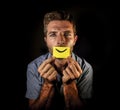 Conceptual dramatic portrait of young depressed man holding paper with happy smiley mouth draw on his lips looking miserable and f Royalty Free Stock Photo