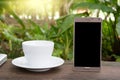 Conceptual green workspace, mobile with blank screen on table, coffee cup and green garden background, business technology conce