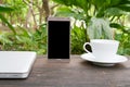 Conceptual green workspace, mobile with blank screen on table, coffee cup and green garden background, business technology conce