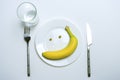 Conceptual image of diet theme. a banana and peas in the form of a smile on a plate, next to a knife and fork on a white table Royalty Free Stock Photo