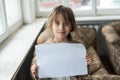 Conceptual image of a sad dejected little girl, holds a blank sheet of paper on gray background. Royalty Free Stock Photo