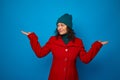 Conceptual portrait of a charming smiling curly haired woman in bright red coat and woolen green hat poses against blue background