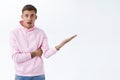 Concerned, frustrated blond young man in pink hoodie, pointing finger right at blank white space, staring camera with