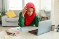 Concerned mature islamic woman using laptop, reading negative news online and touching head, sitting at home