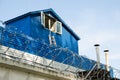 Concrete fence with barbed wire behind which is a white and blue house with an open attic window Royalty Free Stock Photo