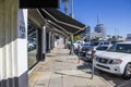 A concrete sidewalk lined with parked meters and parked cars and trucks with lush green palm trees, office buildings and shops