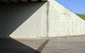 Concrete wall of an underpass divided in two by the shadow of the bridge with grass and sky on th right. Cement sidewalk and road