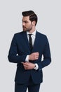 Confidence and charisma. Handsome young man in full suit adjusting sleeve and looking away while standing against grey background