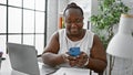 Confident african american business woman, working with laptop and smartphone, smiling boldly at the office interior Royalty Free Stock Photo