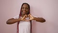 Confident african american woman joyfully forming a heart symbol with her hands, smiling against a lovely isolated pink background