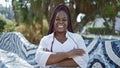 Confident african american woman joyfully stands in city park, arms crossed, sporting a beautiful, relaxed smile