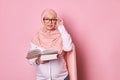Confident Arab Muslim female educator charming pregnant woman in pink hijab and glasses, holding book, looking at camera