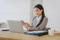 Confident Asian businesswoman using smartphone while working with laptop in office. Royalty Free Stock Photo