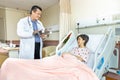 Confident Asian medical doctor explaning to cheerful female patient on medical condition lying on bed in hospital.