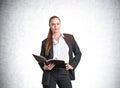 Confident attractive businesswoman in formal suit and shirt is holding a notepad, holding hand on waist and dreaming about future