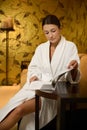 Confident attractive relaxed young woman resting in a lounge zone of a wellness spa clinic, wearing bathrobe, reading magazine and Royalty Free Stock Photo