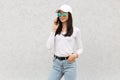 Confident attractive woman wearing baseball cap, casual shirt, jeans and sunglasses, talking via cell phone, dark haired female Royalty Free Stock Photo