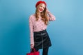 Confident blonde girl in black skirt holding handbag. Studio shot of joyful woman in beret standing on blue background