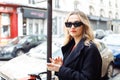 Confident blonde woman wearing sunglasses, black coat while walking in Paris. Lady with red lips hold phone in hand.