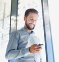 Portrait of smiling businessman texting on smartphone while standing against window in office corridor Royalty Free Stock Photo