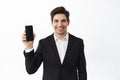 Confident businessman in black suit showing empty smartphone screen, smiling, introduce an application, online store or