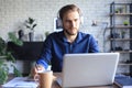 Confident businessman in earphones is writing notes or financial report while sitting at desk with laptop at home Royalty Free Stock Photo