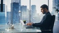 Confident Businessman in a Suit Sitting at a Desk in Modern Office, Using Laptop Computer, Next to Royalty Free Stock Photo