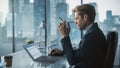 Confident Businessman in a Suit Sitting at a Desk in Modern Office, Using Laptop Computer, Next to Royalty Free Stock Photo