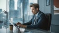 Confident Businessman in a Suit Sitting at a Desk in Modern Office, Using Laptop Computer, Next to Royalty Free Stock Photo