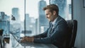 Confident Businessman in a Suit Sitting at a Desk in Modern Office, Using Laptop Computer, Next to Royalty Free Stock Photo