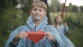 Confident Caucasian boy with paper heart looking back at girl swinging on swings and smiling at camera. Portrait of