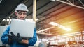 Confident engineer in blue jumpsuit holding laptop computer in a warehouse.