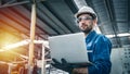 Confident engineer in blue jumpsuit holding laptop computer in a warehouse.