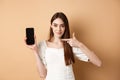 Confident girl pointing at empty smartphone screen, showing online promo deal, standing on beige background